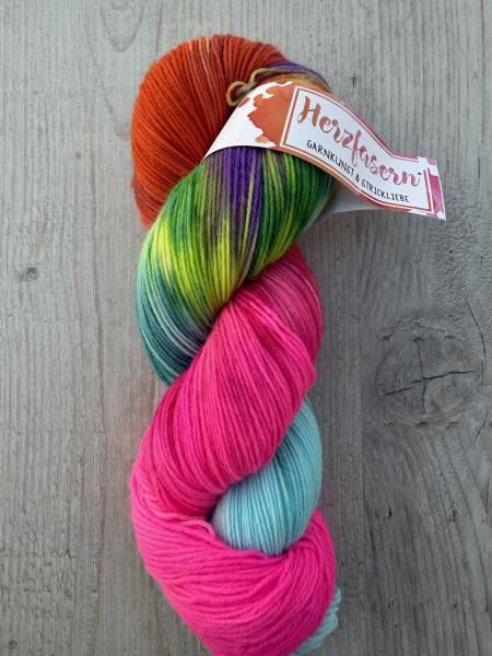 Handgefärbter Wollstrang Regenbogenkuss auf Herzfasern No. 20 mit leuchtendem Pink, Orange, Türkis, Grün, Gelb und violetten Akzenten auf hellem Holzuntergrund.